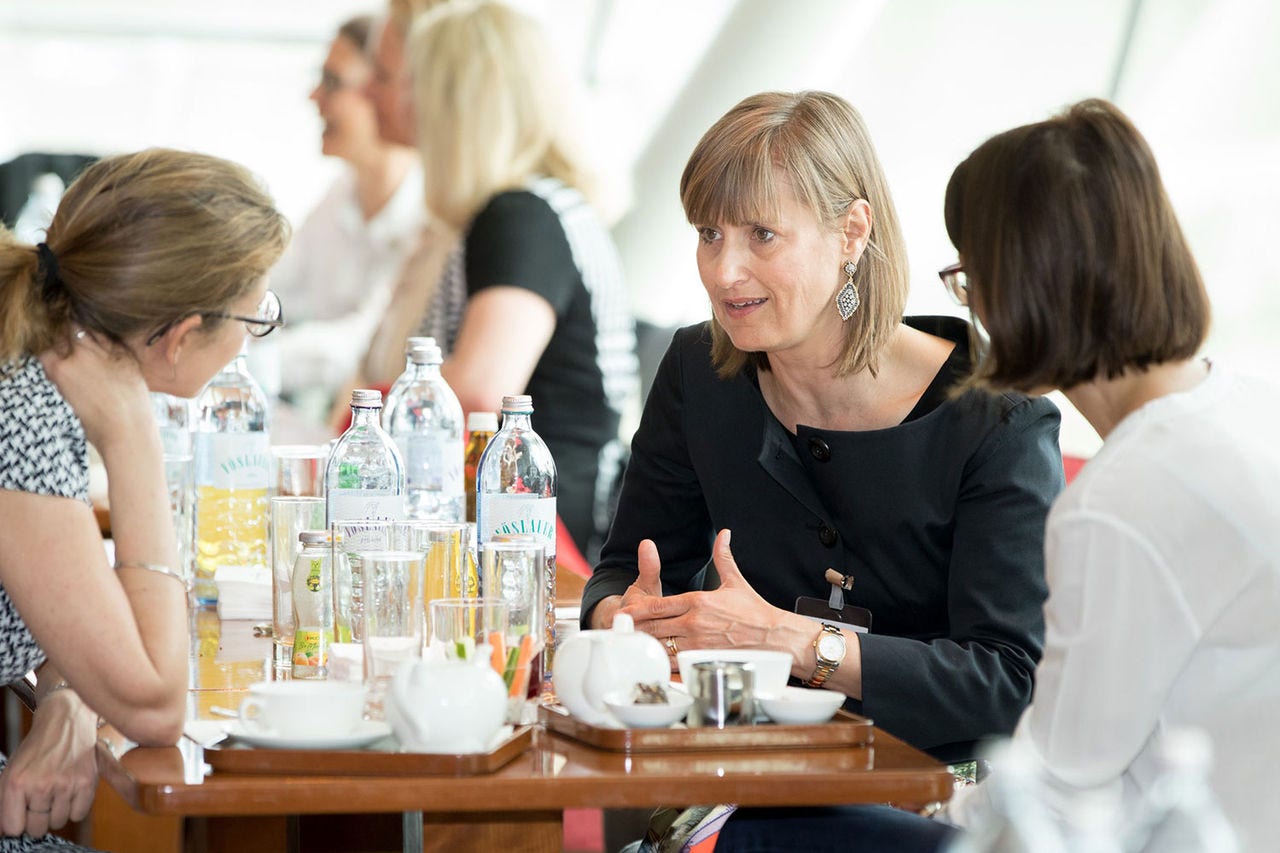 Das Bild zeigt eine Gruppe von Personen, die an einem Tisch sitzen und sich unterhalten. Auf dem Tisch stehen Getränke und Kaffeetassen. Die Umgebung wirkt wie ein Café oder ein informeller Besprechungsraum.