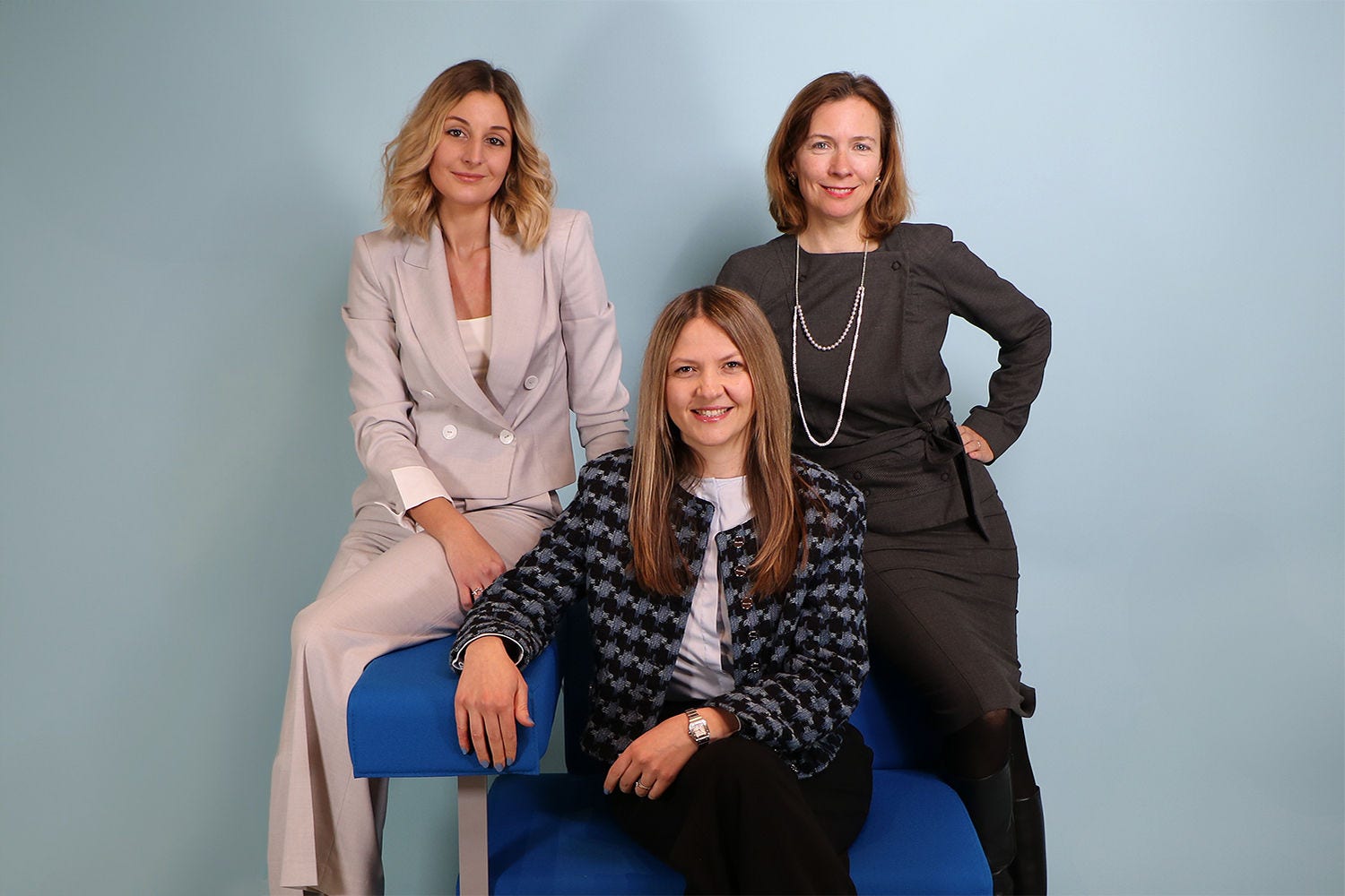 Das Bild zeigt drei Personen in Business-Kleidung vor einem hellblauen Hintergrund, wobei zwei stehen und eine auf einem blauen Stuhl sitzt.