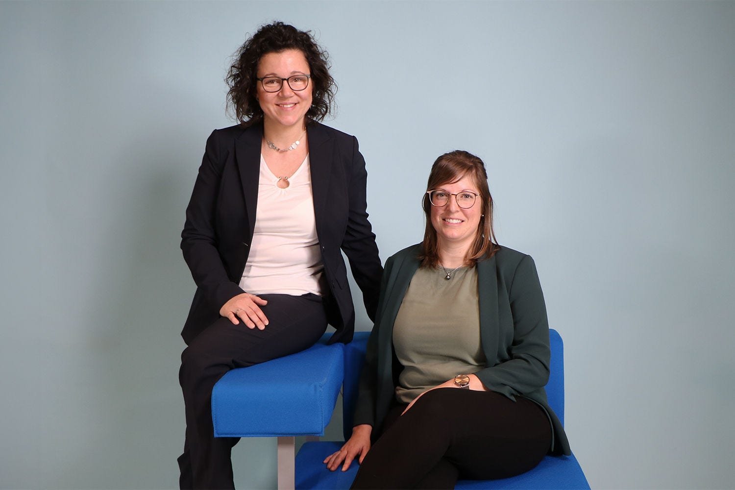 Das Bild zeigt zwei Personen in Business-Kleidung vor einem hellblauen Hintergrund, wobei eine Person auf einem blauen Stuhl sitzt und die andere daneben steht.