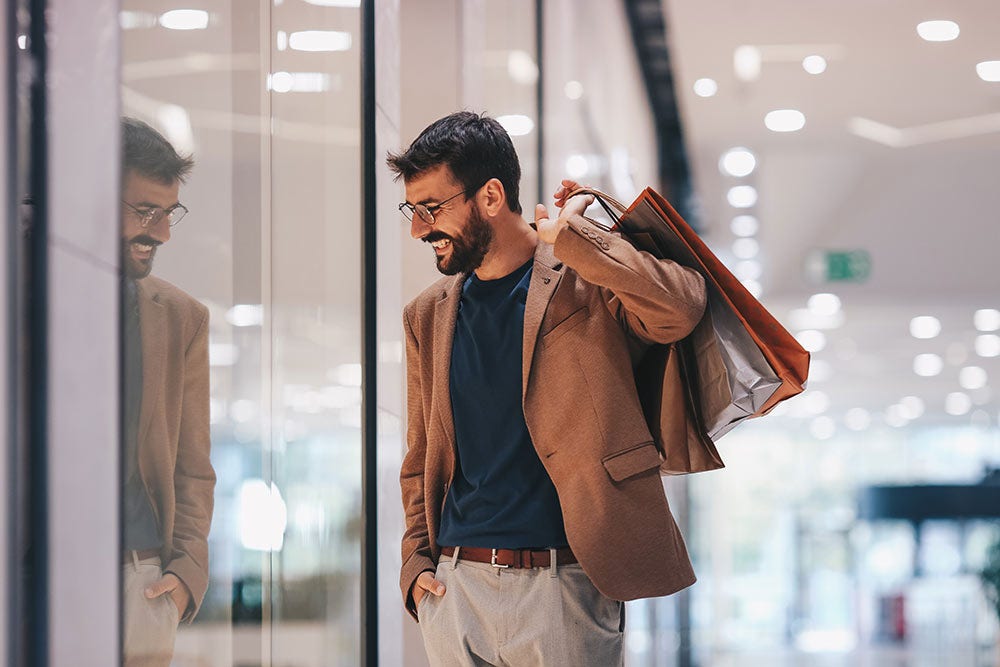 A man walks through a shopping mall with a smile on his face and bags in his hand and looks at the shop windows