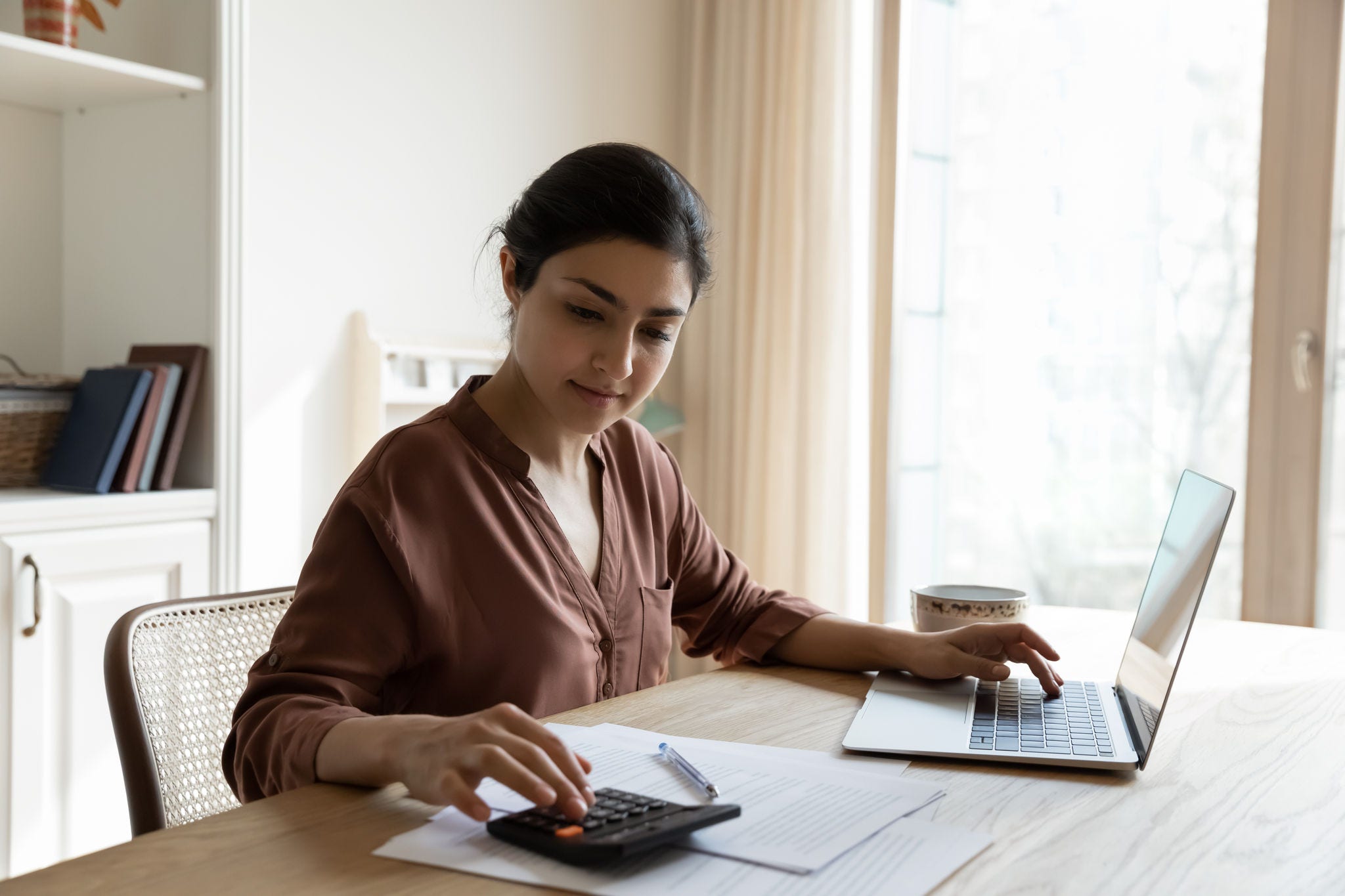 Household budget. Young indian female sit at table calculate utility payments before make money transfer in online bank app on laptop. Focused millennial business woman pay taxes plan personal savings