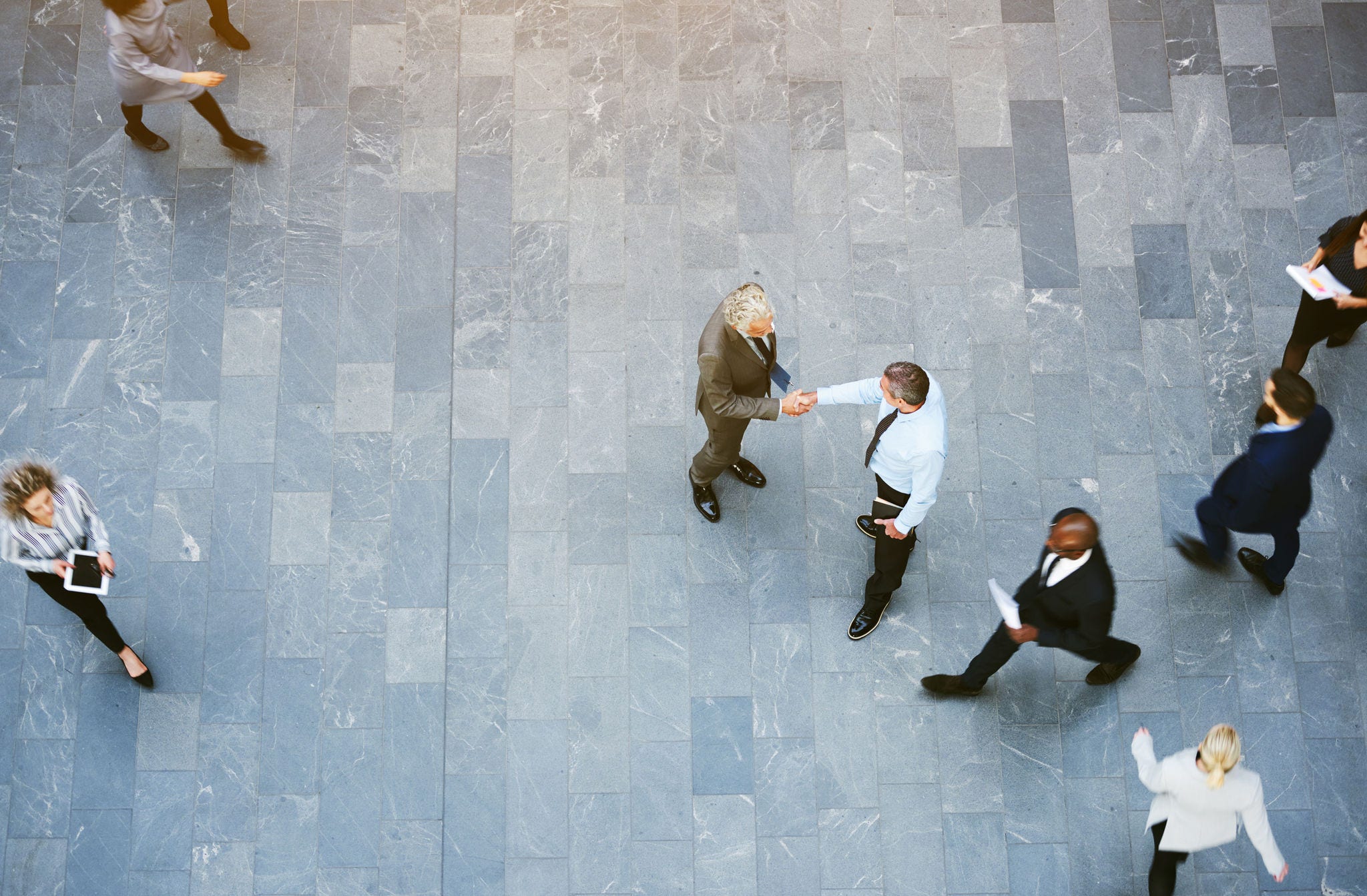 From above two adult business men shaking hands met in office hall with workers walking around.